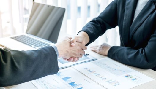 Two people in business attire shake hands at a desk with charts, graphs, and a laptop visible.