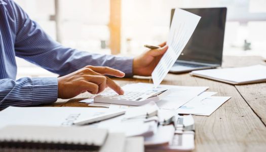 Person in a blue shirt uses a calculator and reviews documents at a desk with a laptop and paperwork.